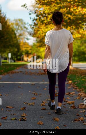 Une jeune femme portant des baskets, un collant violet et un chemisier blanc marche seule sur un sentier de randonnée le jour de l'automne. Le chemin est recouvert de feuilles mortes. Banque D'Images