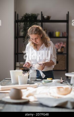 La femelle potter assis et agite la peinture avec un pinceau une tasse sur la table. Femme en céramique. Travail de poterie, compétences artisanales et créatives. Banque D'Images