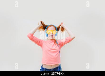 une adolescente avec des cheveux rouges portant des lunettes jaunes et les écouteurs bleus dansent près d'un mur blanc Banque D'Images