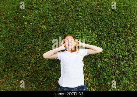 une fille aux cheveux rouges écoute les leçons avec un casque. Couché sur l'herbe Banque D'Images