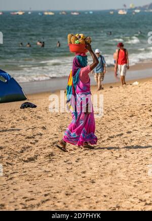 Femme indienne non identifiée en rose coloré bleu sari robe portant des fruits frais dans le bassin rouge à sa tête pour vendre à la rive aux touristes. Ensoleillé Banque D'Images