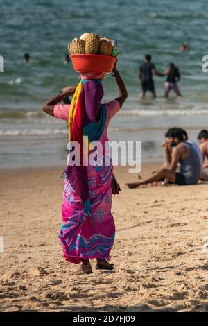 Femme indienne non identifiée en rose coloré bleu sari robe portant des fruits frais dans le bassin rouge à sa tête pour vendre à la rive aux touristes. Ensoleillé Banque D'Images