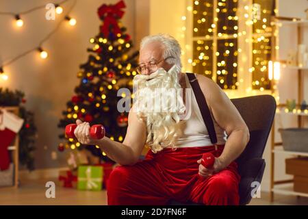 Fort père Noël s'entraîne avec des haltères à la maison sur le fond de l'arbre de Noël. Banque D'Images