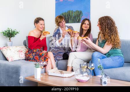 Quatre amies souriantes bavardent et s'asseyant sur le canapé dans le salon tout en mangeant de la pizza, il y a aussi des verres avec du vin sur la table. Banque D'Images