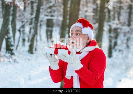 Le Père Noël souffle de la neige. Joyeux Père Noël soufflant sur la neige. Portrait du Père Noël heureux marchant dans une forêt enneigée et soufflant la neige de Noël magique. Banque D'Images