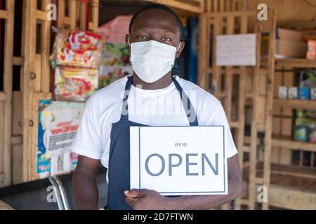 jeune propriétaire de petite entreprise africaine portant un masque facial et en tenant une affiche ouverte devant son magasin Banque D'Images