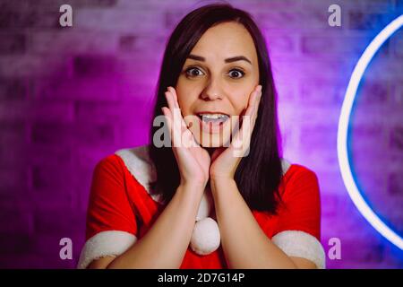 Une jeune femme en costume du Père Noël ouvre sa bouche par surprise sur fond de mur éclairé. Portrait de brunette en costume de Noël avec les mains sur Banque D'Images