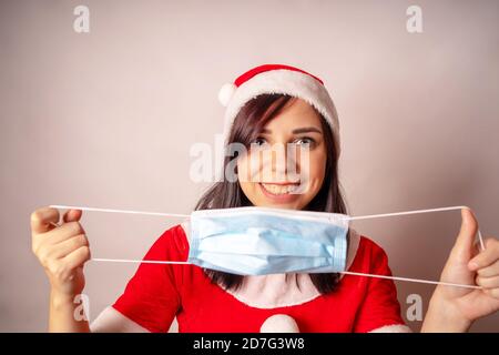 Femme en costume de Père Noël avec masque médical dans les mains sur fond gris. Portrait de brunette en chapeau de Noël tenant le masque de protection. Concept de Banque D'Images