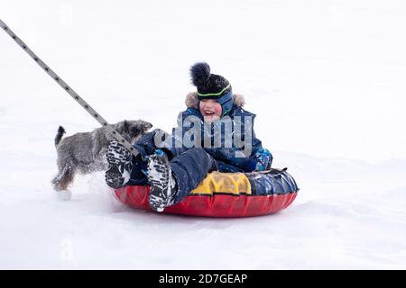 Un garçon en vêtements d'hiver fait un traîneau gonflable sur la neige blanche, un chien joue avec un garçon. Concept de jeux d'hiver en plein air. Banque D'Images