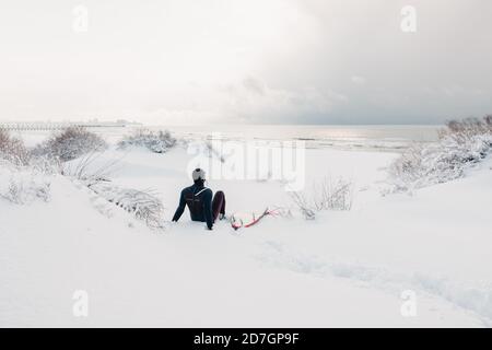 Hiver froid et surfeur s'assoient sur la plage enneigée avec surf et voir aux vagues. Jour d'hiver avec surfeur en combinaison. Banque D'Images