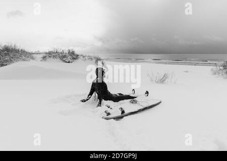 Hiver froid et surfeur s'assoient sur la plage enneigée avec surf et voir aux vagues. Jour d'hiver avec surfeur en combinaison. Banque D'Images