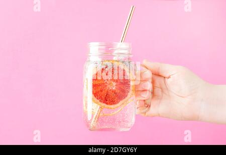 Main femelle tenant une boisson d'été avec soda, tranche d'orange et graines de chia dans un pot de verre sur fond rose, foyer sélectif Banque D'Images