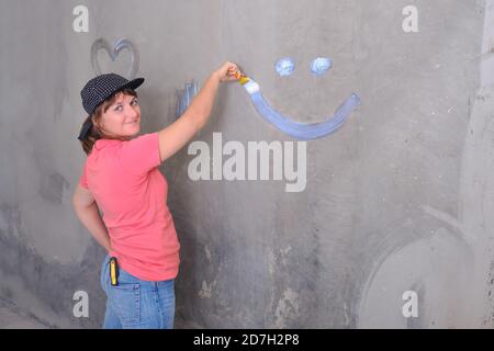 Le peintre peint le mur avec de la peinture blanche. Réparer dans une maison neuve par eux-mêmes. Une femme dans un chapeau dessine un sourire sur le mur, l'espace de copie. Banque D'Images