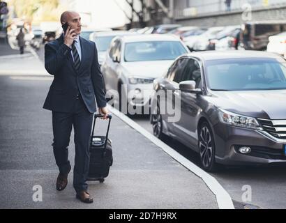 Homme d'affaires parle sur téléphone portable tenant une valise à la main. Homme caucasien d'âge moyen marchant le long du trottoir devant les voitures garées. Concept de voyage d'affaires Banque D'Images