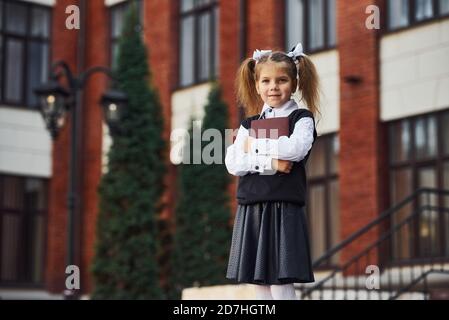 Jeune fille d'école en tenue formelle et bloc-notes dans les mains stands près du bâtiment extérieur Banque D'Images