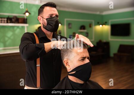 dans un salon de coiffure, un coiffeur peigne un grand homme avec sa main et son peigne, il porte des masques pour la prévention du coronavirus pandémique. le client est assis. hanches Banque D'Images