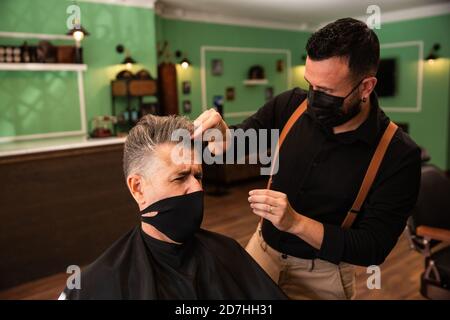 dans un salon de coiffure, un coiffeur peigne un grand homme avec sa main et son peigne, il porte des masques pour la prévention du coronavirus pandémique. le client est assis. hanches Banque D'Images