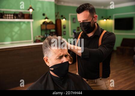 dans un salon de coiffure, un coiffeur peigne un grand homme avec sa main et son peigne, il porte des masques pour la prévention du coronavirus pandémique. le client est assis. hanches Banque D'Images