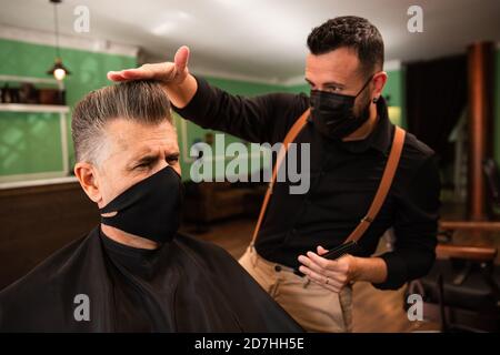 dans un salon de coiffure, un coiffeur peigne un grand homme avec sa main et son peigne, il porte des masques pour la prévention du coronavirus pandémique. le client est assis. hanches Banque D'Images