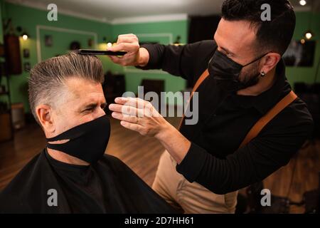 dans un salon de coiffure, un coiffeur peigne un grand homme avec sa main et son peigne, il porte des masques pour la prévention du coronavirus pandémique. le client est assis. hanches Banque D'Images