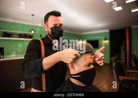 dans un salon de coiffure, un coiffeur peigne un grand homme avec sa main et son peigne, il porte des masques pour la prévention du coronavirus pandémique. le client est assis. hanches Banque D'Images