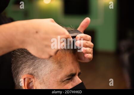 dans un salon de coiffure, un coiffeur peigne un grand homme avec sa main et son peigne, il porte des masques pour la prévention du coronavirus pandémique. le client est assis. hanches Banque D'Images