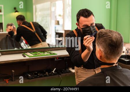 dans un salon de coiffure, un coiffeur peigne un grand homme avec sa main et son peigne, il porte des masques pour la prévention du coronavirus pandémique. le client est assis. hanches Banque D'Images