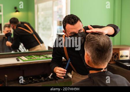 dans un salon de coiffure, un coiffeur peigne un grand homme avec sa main et son peigne, il porte des masques pour la prévention du coronavirus pandémique. le client est assis. hanches Banque D'Images