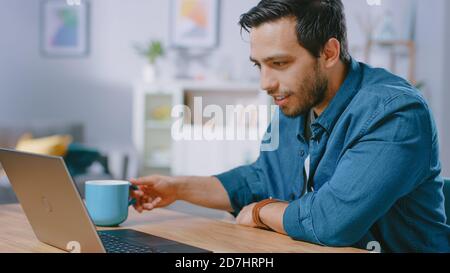 Portrait d'un jeune homme souriant travaillant sur un ordinateur portable à la maison et prenant sa tasse de café. Il est satisfait de son travail ou de son film. Confortable et moderne plat fait Banque D'Images