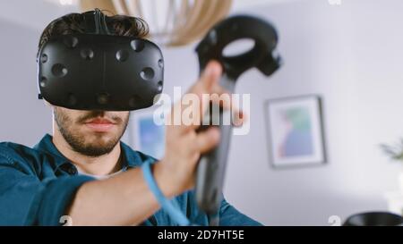 Un jeune homme énergique portant un casque de réalité virtuelle et des contrôleurs joue dans un jeu vidéo à la maison. Homme actif jouant VR Quest Adventure Banque D'Images