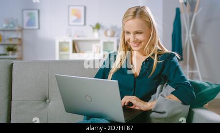 Belle jeune femme se détend sur un canapé à la maison, utilise un ordinateur portable, navigue par Internet, regarde des vidéos amusantes et fait e-Shopping. Banque D'Images