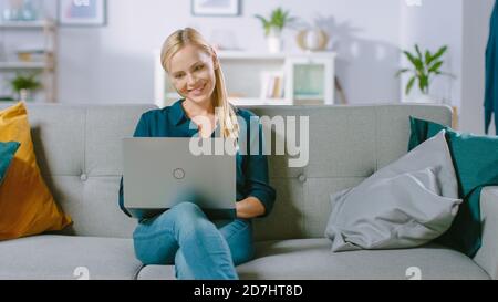 Belle jeune femme se détend sur un canapé à la maison, utilise un ordinateur portable, navigue par Internet, regarde des vidéos amusantes et fait e-Shopping. Banque D'Images