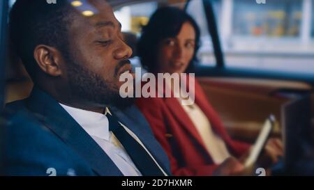 Jeune femme et homme à cheval sur le siège arrière d'une voiture dans la soirée. L'homme utilise un smartphone. Prise de vue réalisée à l'extérieur de la voiture. Banque D'Images
