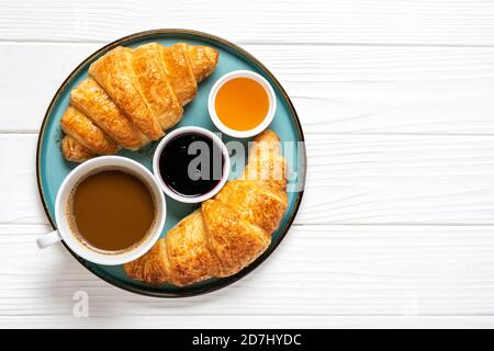 Croissants français frais avec chocolat sur l'assiette, confiture de myrtilles, miel, tasse de café blanc sur fond de bois blanc petit-déjeuner continental le matin Banque D'Images