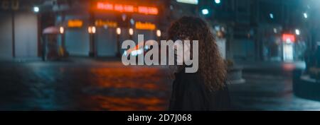 Femme aux cheveux bouclés dans une chemise blanche et noire la veste descend la nuit dans la rue rouge Et tourne et regarde la caméra arrière - mode Banque D'Images