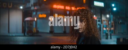 Femme aux cheveux bouclés dans une chemise blanche et noire la veste descend la nuit dans la rue rouge Et tourne et regarde la caméra arrière - mode Banque D'Images