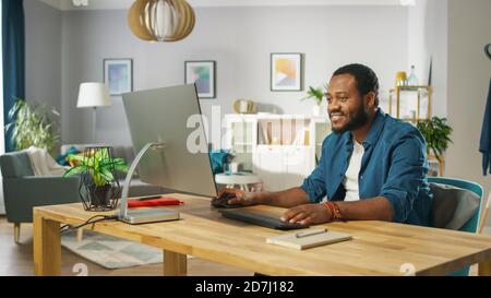 Beau Black Man travaille sur un ordinateur personnel tout en étant assis au bureau de son salon confortable. Banque D'Images