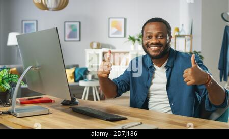 Portrait de l'homme noir beau travaille sur un ordinateur personnel, a le succès et montre Thumbs Up avec les deux mains. Dans le salon confortable en arrière-plan. Banque D'Images