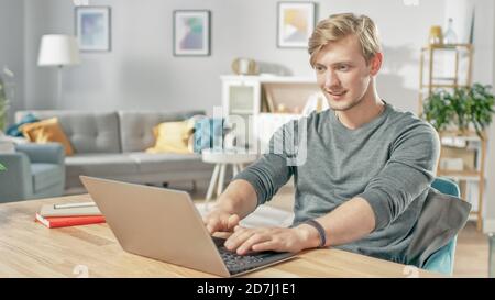 Beau jeune homme souriant utilisant un ordinateur portable tout en étant assis au bureau de son salon confortable. Banque D'Images