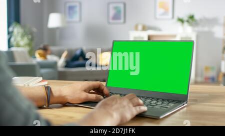 Prise de vue au-dessus de l'épaule des mains du Mans en tapant sur un ordinateur portable avec écran vert Mock-up. En arrière-plan salon confortable avec femme se reposant sur un canapé. Banque D'Images