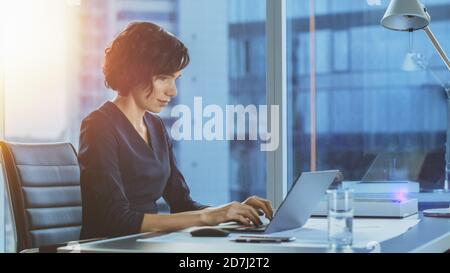 Vue latérale Portrait de la belle femme d'affaires travaillant sur un ordinateur portable dans son bureau moderne avec vue fenêtre CityScape. Une femme exécutive utilise un ordinateur. Banque D'Images