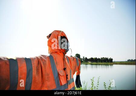 Un vrai sourcil contre un ciel bleu portant un costume de travail orange Et gants - Scarecrow avec visage effrayant devant Le lac - Halloween concept et la sécurité Banque D'Images