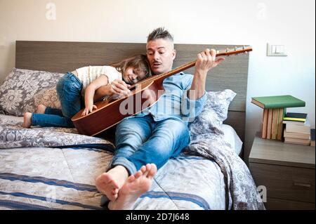 Cool Daddy jouer de la guitare acoustique et enseigner une chanson Pendant que sa fille l'écoute - papa et fille sont assis sur le lit jouant de la guitare Banque D'Images