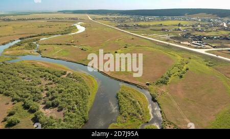 Un drone survole une rivière de couleur bleue entourée de forêts et de champs avec une vue aérienne sur la route terrestre et la silhouette du véhicule de conduite. Village local Banque D'Images