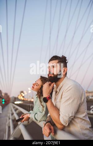 Un couple attentionné qui regarde loin tout en s'inclinant sur la rambarde au pont en ville au coucher du soleil Banque D'Images