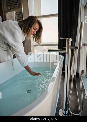 Femme âgée se pliant tout en examinant la température dans la baignoire à hôtel de luxe Banque D'Images