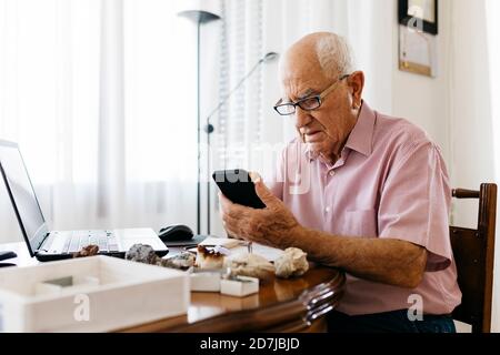 Homme senior utilisant un smartphone tout en faisant des recherches sur les minéraux et fossile à la maison Banque D'Images