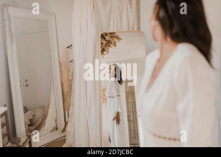 Jeune mariée regardant dans le miroir tout en se tenant à la robe de mariage boutique Banque D'Images