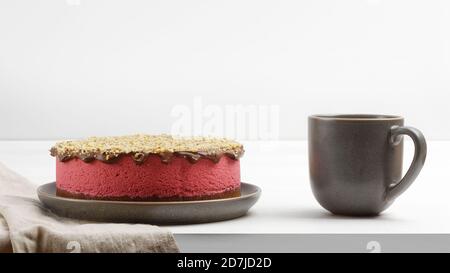 Cheesecake maison à la framboise parsemé de chapelure de noix et d'une tasse de thé sur une table en bois blanc. Vue avant. Banque D'Images