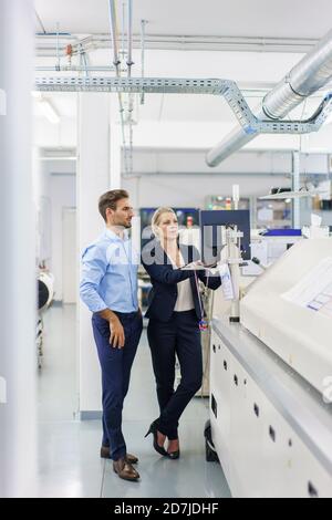 Des collègues hommes et femmes discutent en regardant l'ordinateur sur machines dans une usine éclairée Banque D'Images